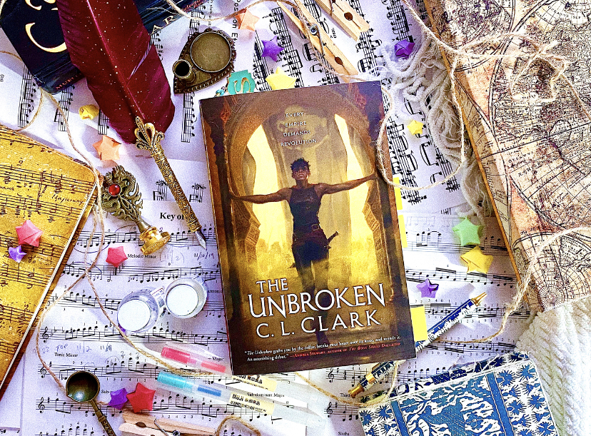 A flat lay of The Unbroken by C. L. Clark atop violin sheet music and a white woven blanket. To the left of the book is a quill pen, and to the right is a box displaying a map of the world.